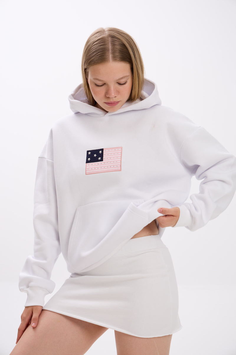Woman wearing a white hoodie with a pink and navy flag patch detail on the chest, paired with a white mini skirt. She is looking down and slightly lifting the hem of the hoodie, showing a casual styling detail against a plain white background.