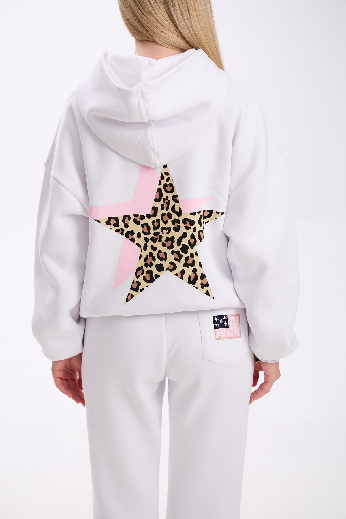 Back view of a woman wearing a white hoodie with a large leopard print star design over a pink star on the back, paired with matching white sweatpants featuring a small patch detail on the pocket, against a plain background