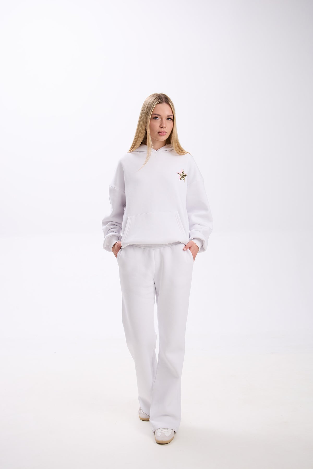 Woman wearing a white hoodie with a small leopard print star and pink accent on the chest, paired with matching white sweatpants and white sneakers, standing against a plain background