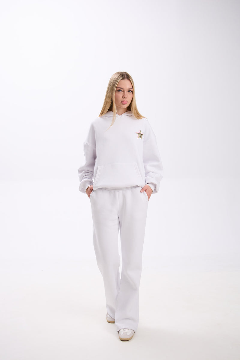Woman wearing a white hoodie with a small leopard print star and pink accent on the chest, paired with matching white sweatpants and white sneakers, standing against a plain background