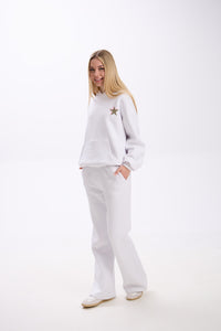 Woman wearing a white hoodie with a small leopard print star and pink accent on the chest, paired with matching white sweatpants and white sneakers, smiling with hands in pockets on a plain background