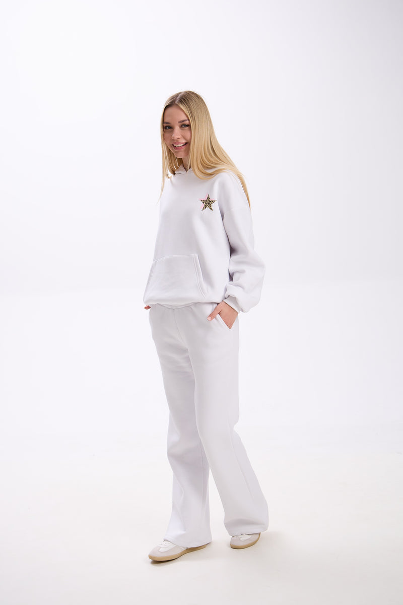 Woman wearing a white hoodie with a small leopard print star and pink accent on the chest, paired with matching white sweatpants and white sneakers, smiling with hands in pockets on a plain background