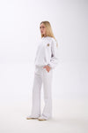 Side view of a woman wearing a white hoodie with a small leopard print star and pink accent on the chest, paired with matching white wide-leg sweatpants and white sneakers, against a plain background