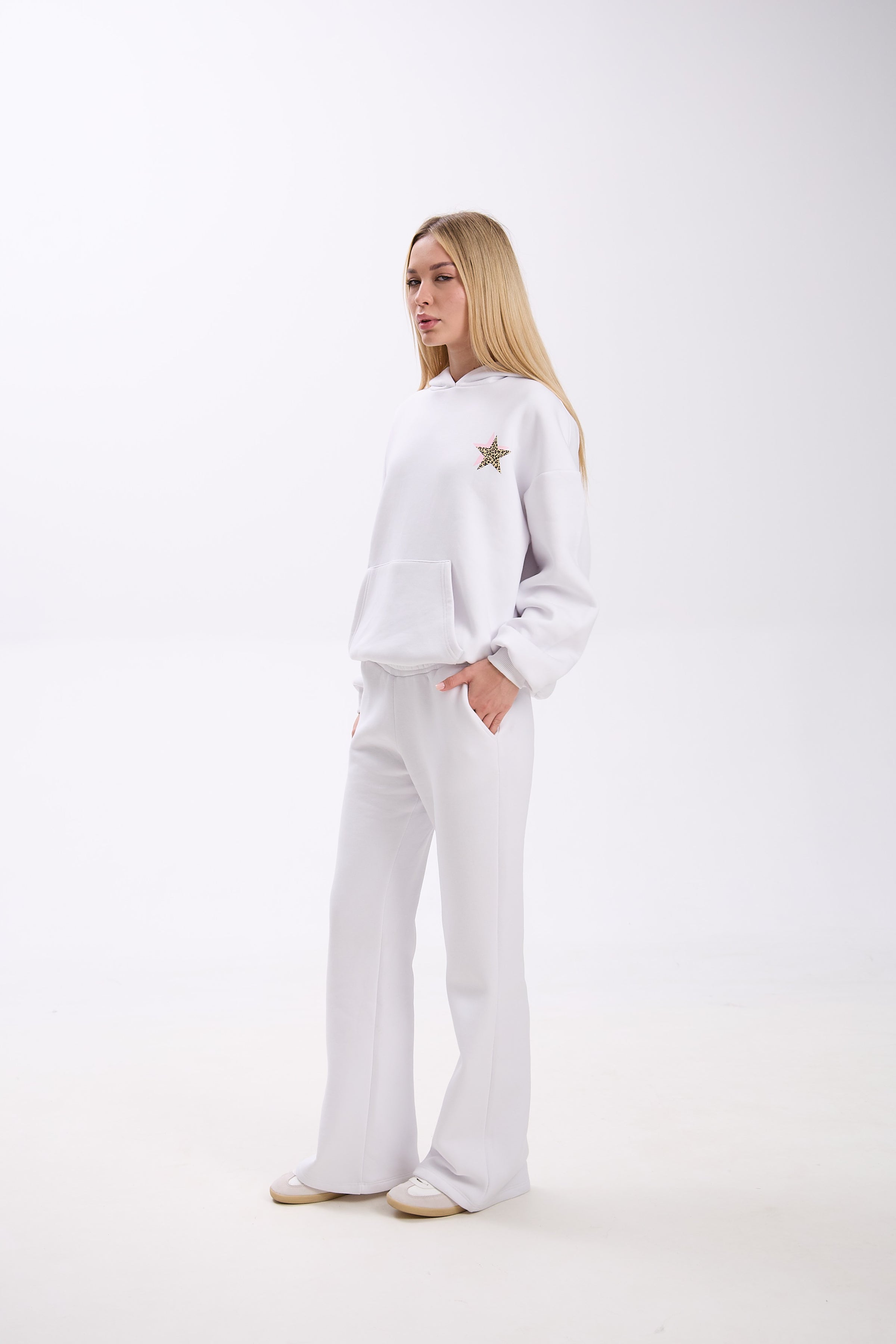 Side view of a woman wearing a white hoodie with a small leopard print star and pink accent on the chest, paired with matching white wide-leg sweatpants and white sneakers, against a plain background