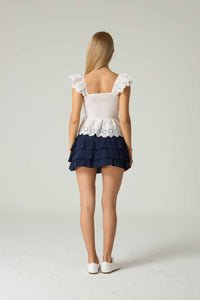 Woman wearing a white top with lace details and a navy blue skirt on a light gray background.