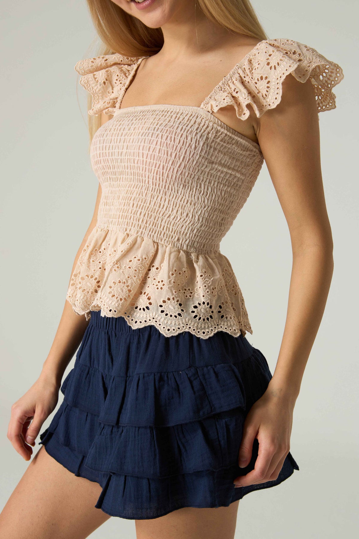 Woman wearing a beige top with lace details and a navy blue skirt on a plain background.
