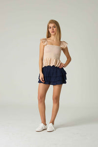 Woman wearing a beige top with ruffled details and a navy blue skirt on a white background.