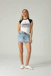 Person wearing a white and black baseball shirt with '1990 England' text, denim shorts, and white sneakers on a plain background.