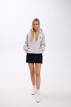 Woman wearing a gray hoodie with a pink hood, navy skirt, and white sneakers on a white background