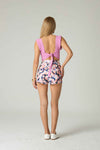Woman wearing a pink top with open back and floral shorts on a white background.
