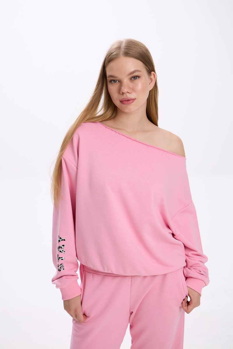 Model in a pink off-shoulder sweatshirt with logo detail on the sleeve, paired with matching pink sweatpants.