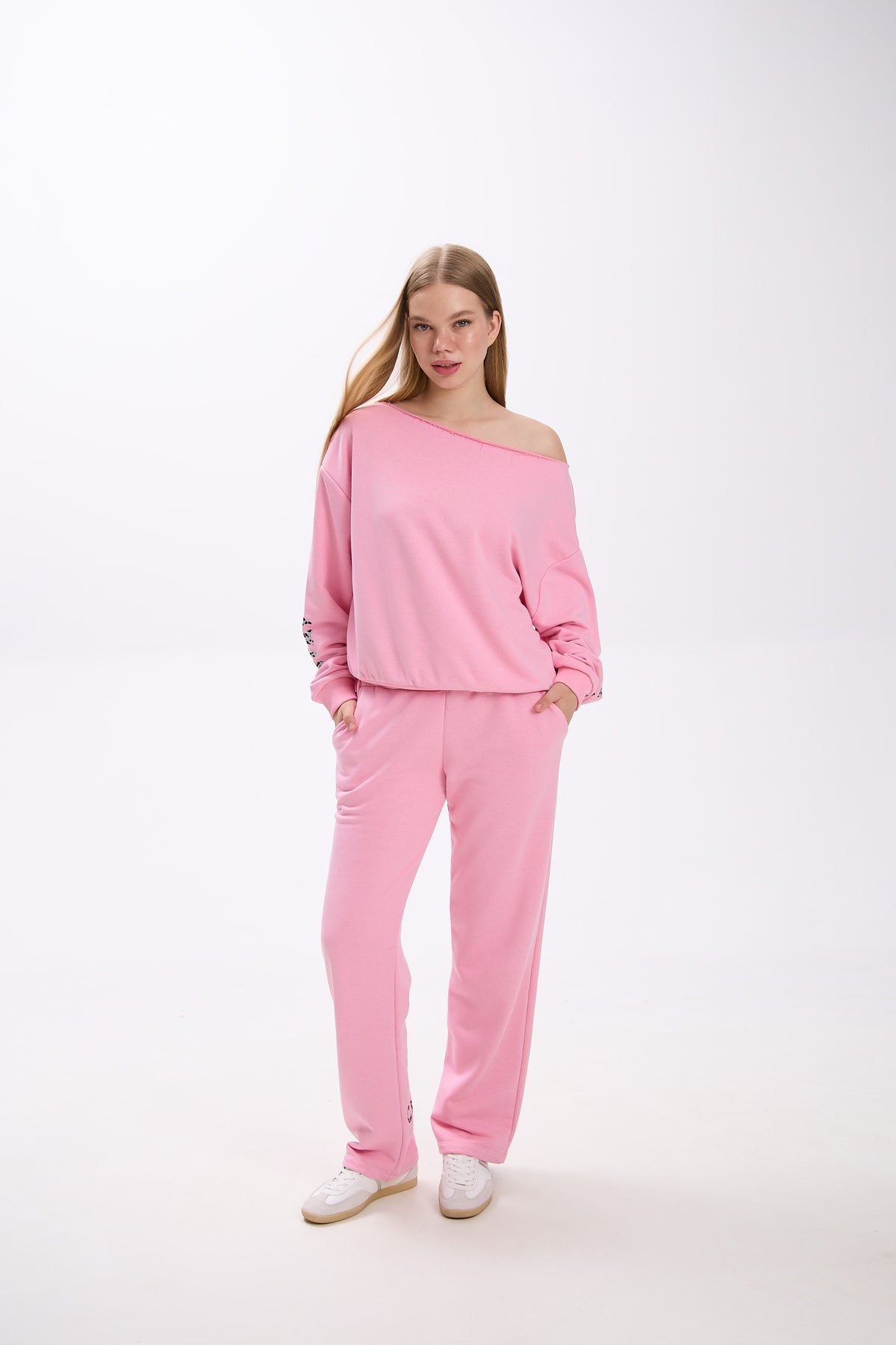 Model in a pink off-shoulder sweatshirt with logo sleeve detail, paired with matching pink sweatpants and sneakers.