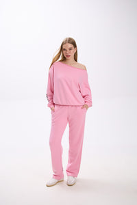 Model in a pink off-shoulder sweatshirt with logo sleeve detail, paired with matching pink sweatpants and sneakers.