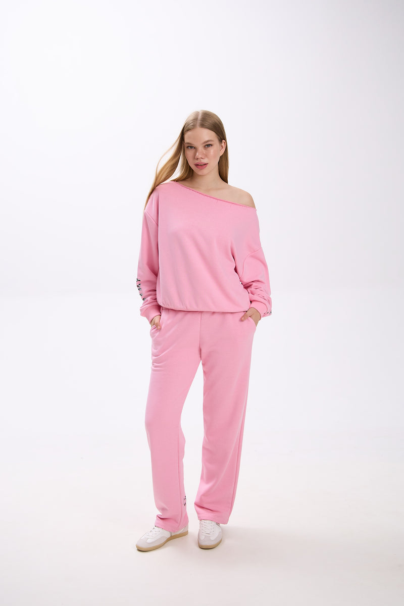 Model in a pink off-shoulder sweatshirt with logo sleeve detail, paired with matching pink sweatpants and sneakers.