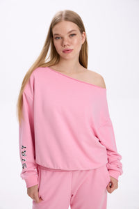 Model in a pink off-shoulder sweatshirt with logo detail on the sleeve, paired with matching pink sweatpants.