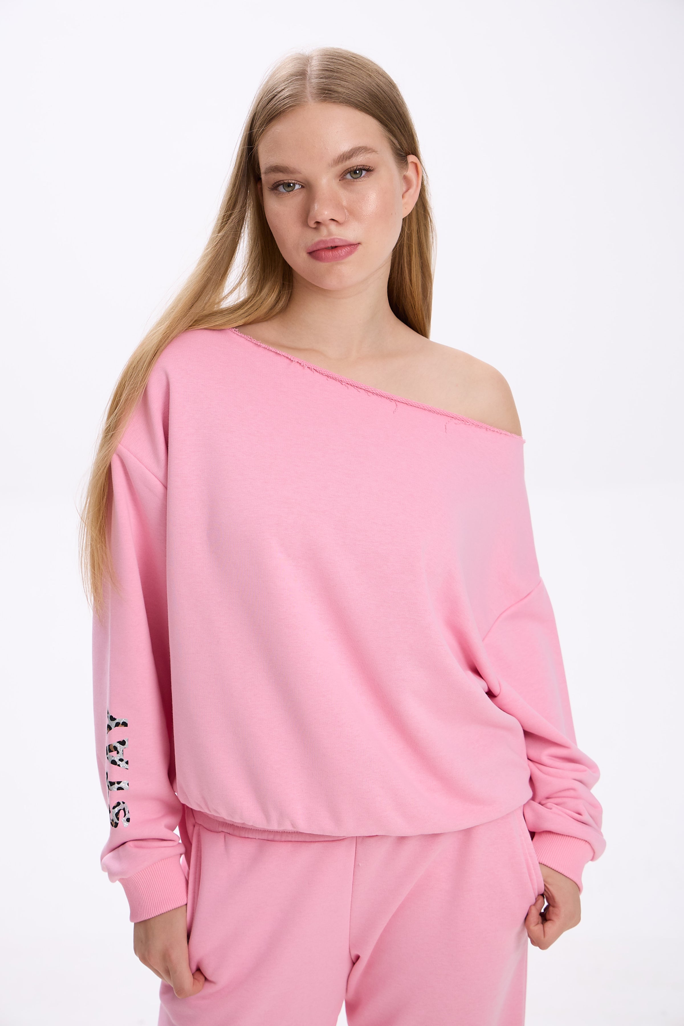 Model in a pink off-shoulder sweatshirt with logo detail on the sleeve, paired with matching pink sweatpants.