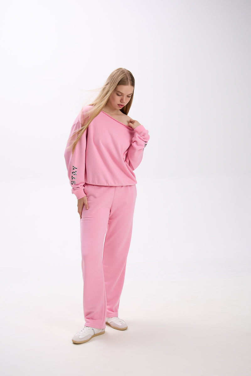 Model in a pink off-shoulder sweatshirt with sleeve logo detail, styled with matching sweatpants and white sneakers.