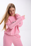 Model in a pink off-shoulder sweatshirt with logo sleeve detail and matching sweatpants, posing in motion.