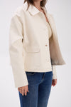 Person wearing a beige jacket over a white shirt with blue jeans on a white background.