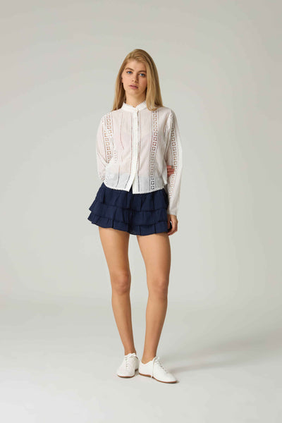 Woman wearing a white lace blouse and navy skirt on a plain background.