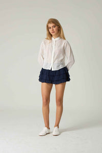 Woman wearing a white lace blouse and navy shorts on a plain background.