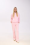 A woman wearing a light pink hoodie paired with matching wide-leg sweatpants and white sneakers, posing with hands in pockets against a plain white background.