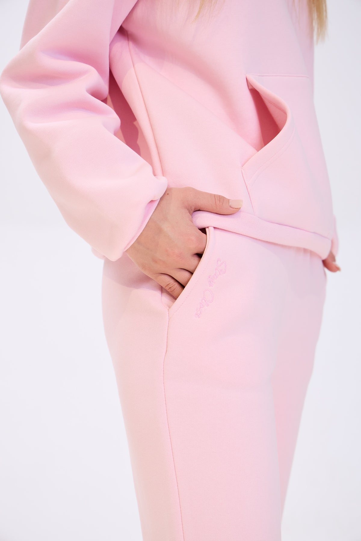 Close-up of a woman wearing light pink sweatpants with her hand in the side pocket, paired with a matching hoodie, highlighting the fabric texture and pocket detail.