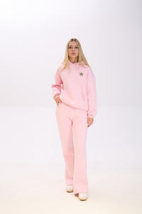 Woman wearing a light pink hoodie with a star patch on the chest, paired with matching pink sweatpants and white sneakers, posing with one hand in her pocket against a plain white background.