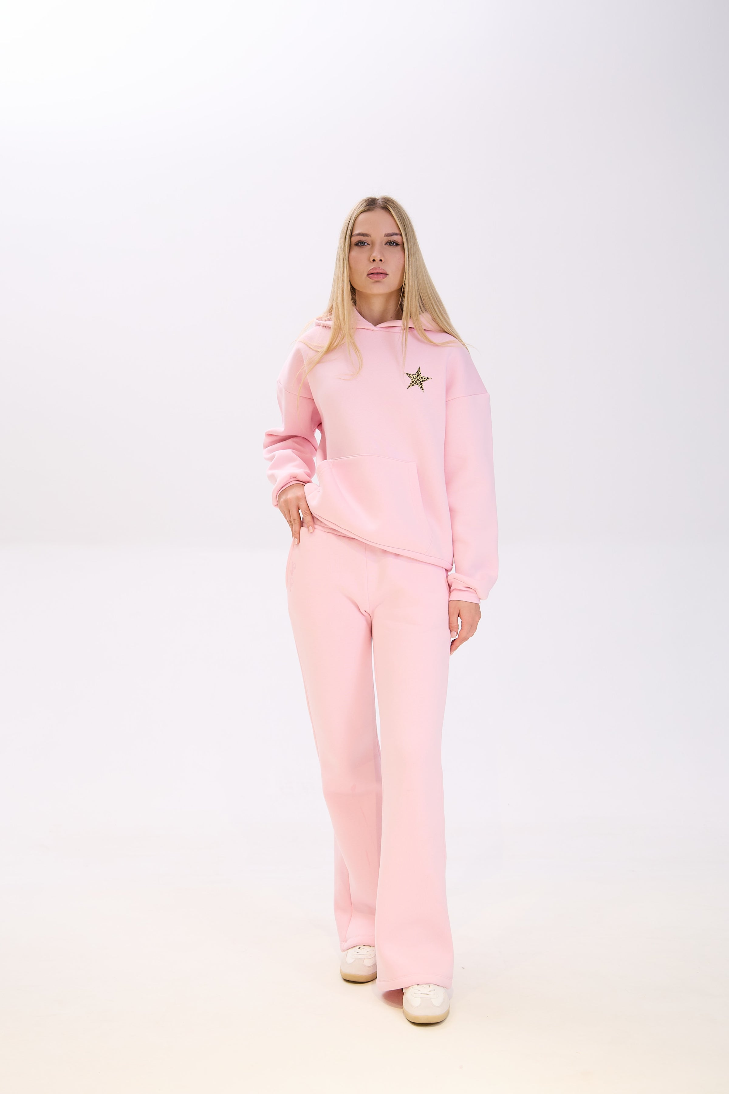 Woman wearing a light pink hoodie with a star patch on the chest, paired with matching pink sweatpants and white sneakers, posing with one hand in her pocket against a plain white background.