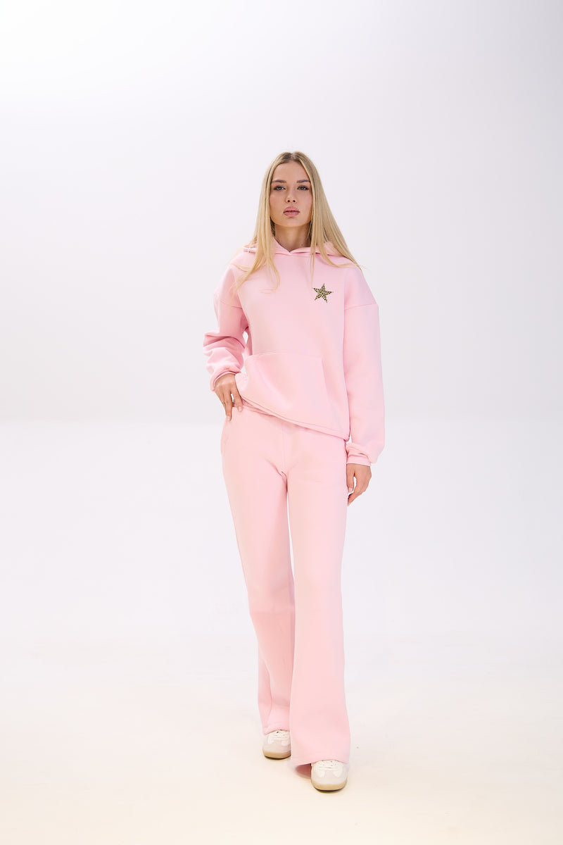 Woman wearing a light pink hoodie with a star patch on the chest, paired with matching pink sweatpants and white sneakers, posing with one hand in her pocket against a plain white background.