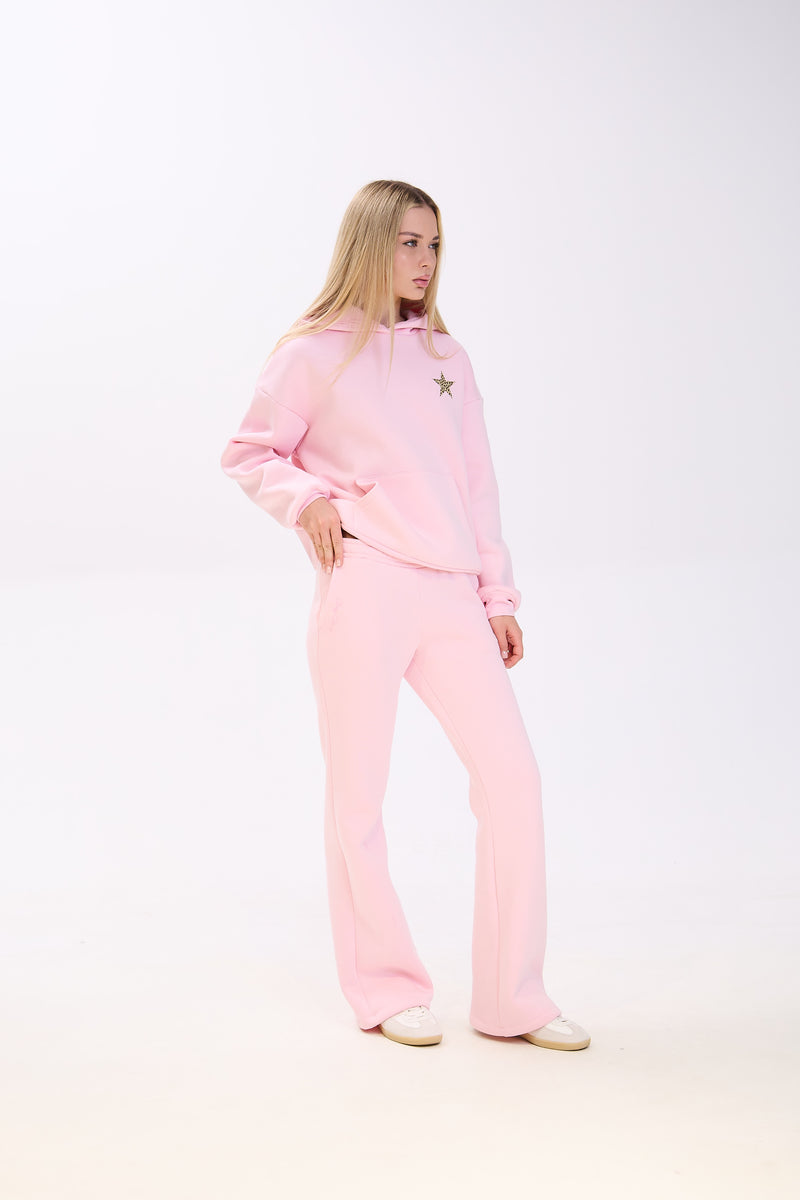 Woman posing in a light pink hoodie with a small star patch on the chest, paired with matching pink flared sweatpants and white sneakers, standing against a plain white background.