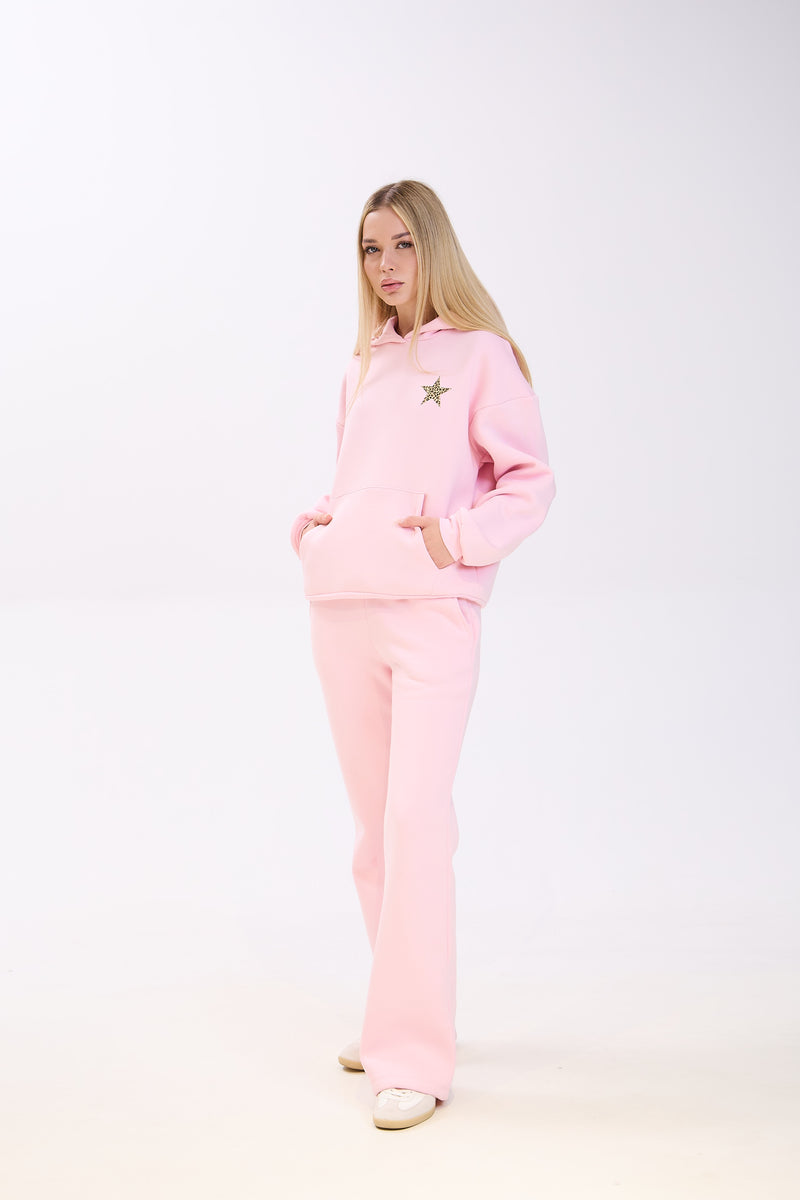 Woman modeling a pastel pink hoodie with a small star patch on the chest, paired with matching pink flared sweatpants and white sneakers, posing with hands in pockets against a plain white background.