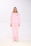 Woman wearing a light pink hoodie with a small star patch on the chest, paired with matching pink sweatpants and white sneakers, standing straight against a plain white background.