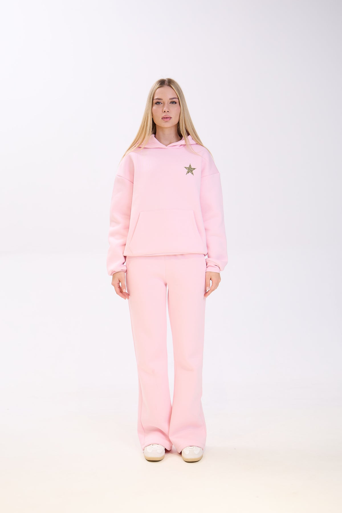 Woman wearing a light pink hoodie with a small star patch on the chest, paired with matching pink sweatpants and white sneakers, standing straight against a plain white background.