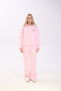 Woman wearing a light pink hoodie with a small star patch on the chest, paired with matching pink sweatpants and white sneakers, standing straight against a plain white background.