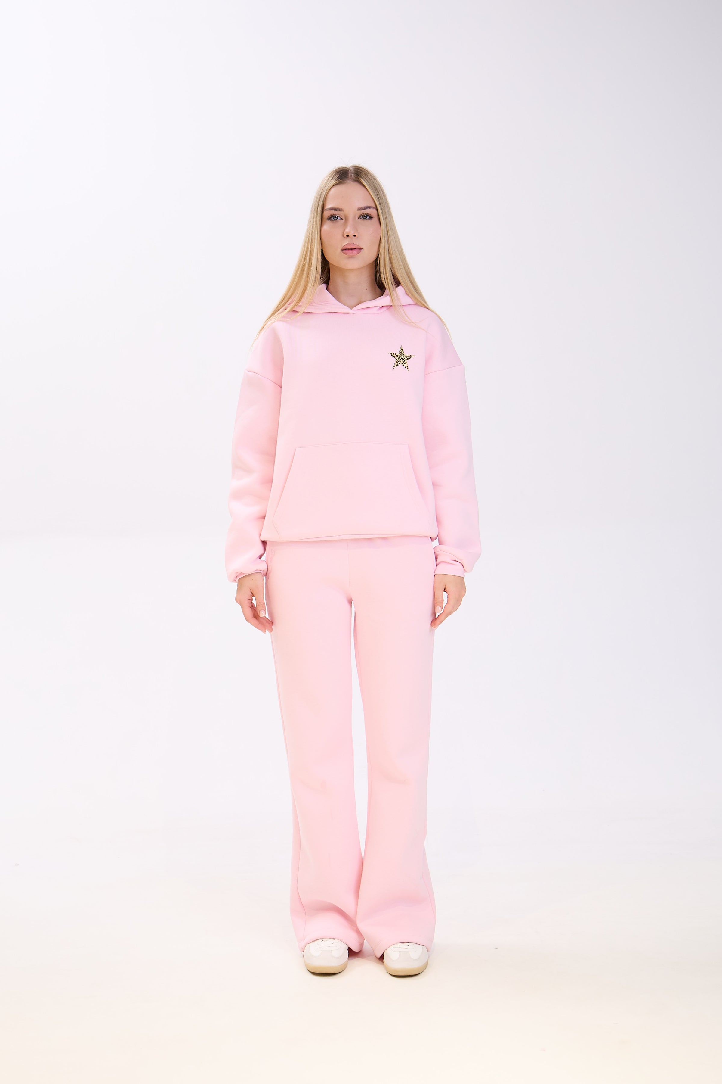 Woman wearing a light pink hoodie with a small star patch on the chest, paired with matching pink sweatpants and white sneakers, standing straight against a plain white background.