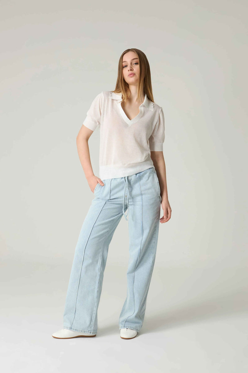 Woman wearing a light-colored blouse and light blue pants against a plain background.