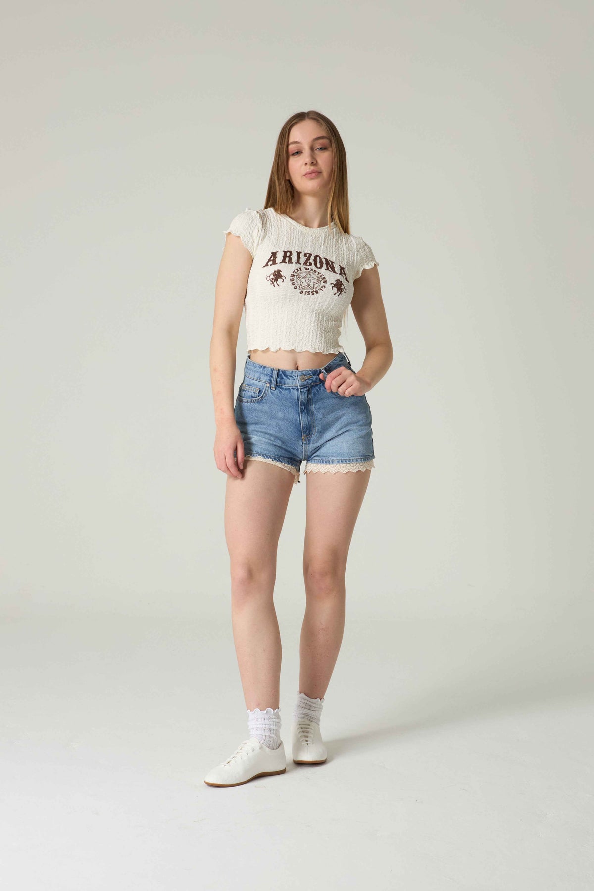 Person wearing a white crop top with 'Arizona' text and denim shorts on a plain background.