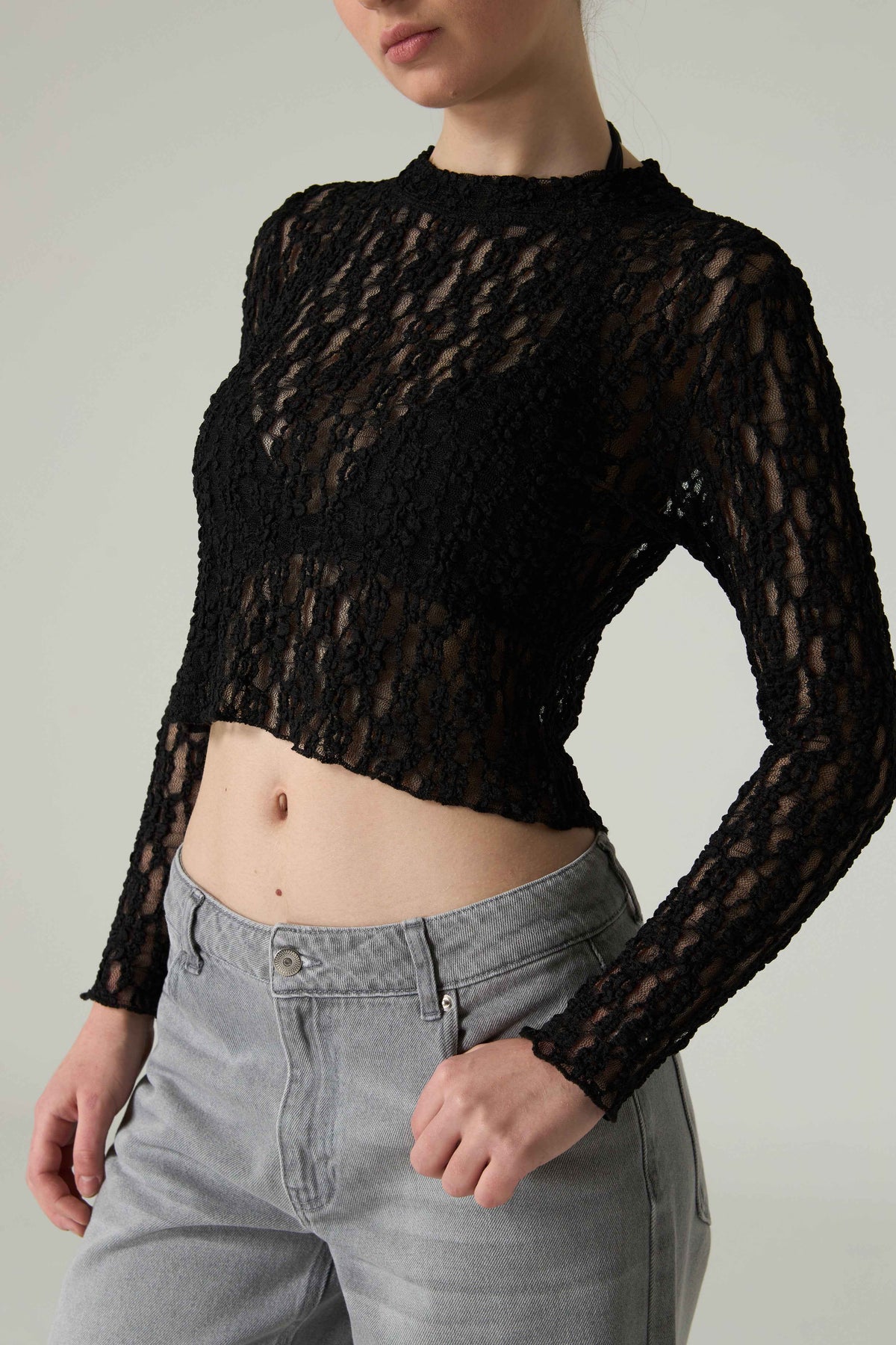 Person wearing a black sheer long-sleeve top and gray pants on a plain background.