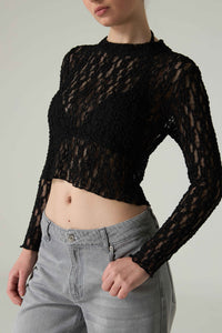 Person wearing a black sheer long-sleeve top and gray pants on a plain background.