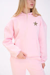 Close-up of a woman wearing a pastel pink hoodie with a leopard print star detail on the chest, paired with matching pink sweatpants, shown against a plain white background.