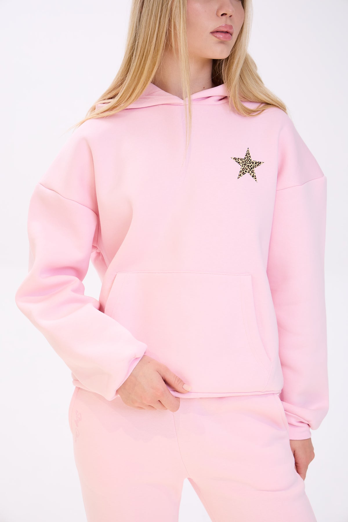 Close-up of a woman wearing a pastel pink hoodie with a leopard print star detail on the chest, paired with matching pink sweatpants, shown against a plain white background.