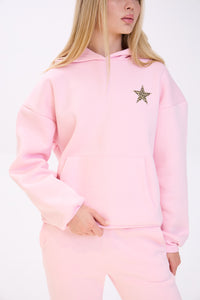 Close-up of a woman wearing a pastel pink hoodie with a leopard print star detail on the chest, paired with matching pink sweatpants, shown against a plain white background.
