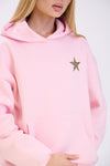 Close-up of a woman in a pastel pink hoodie featuring a leopard print star detail on the chest, shown with a kangaroo pocket and minimal styling against a plain white background.
