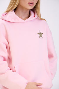 Close-up of a woman in a pastel pink hoodie featuring a leopard print star detail on the chest, shown with a kangaroo pocket and minimal styling against a plain white background.