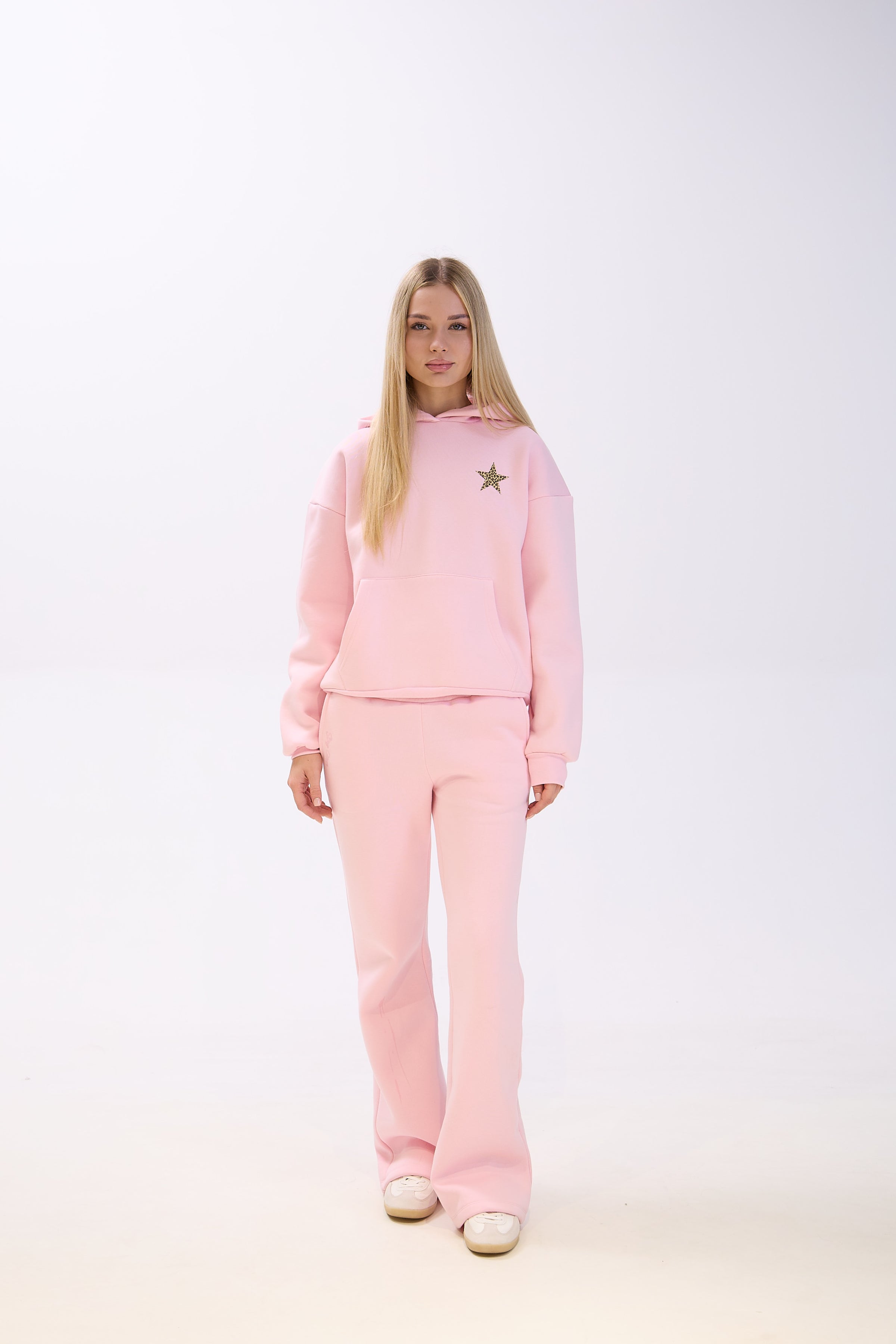 Woman wearing a light pink hoodie with a leopard print star detail on the chest, paired with matching wide-leg pink pants and white sneakers, standing against a plain white background.