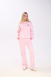 Woman dressed in a pastel pink hoodie with a leopard print star detail and matching pink wide-leg pants, posing with her hands in the pockets against a white studio background.