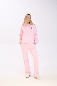 Woman dressed in a pastel pink hoodie with a leopard print star detail and matching pink wide-leg pants, posing with her hands in the pockets against a white studio background.