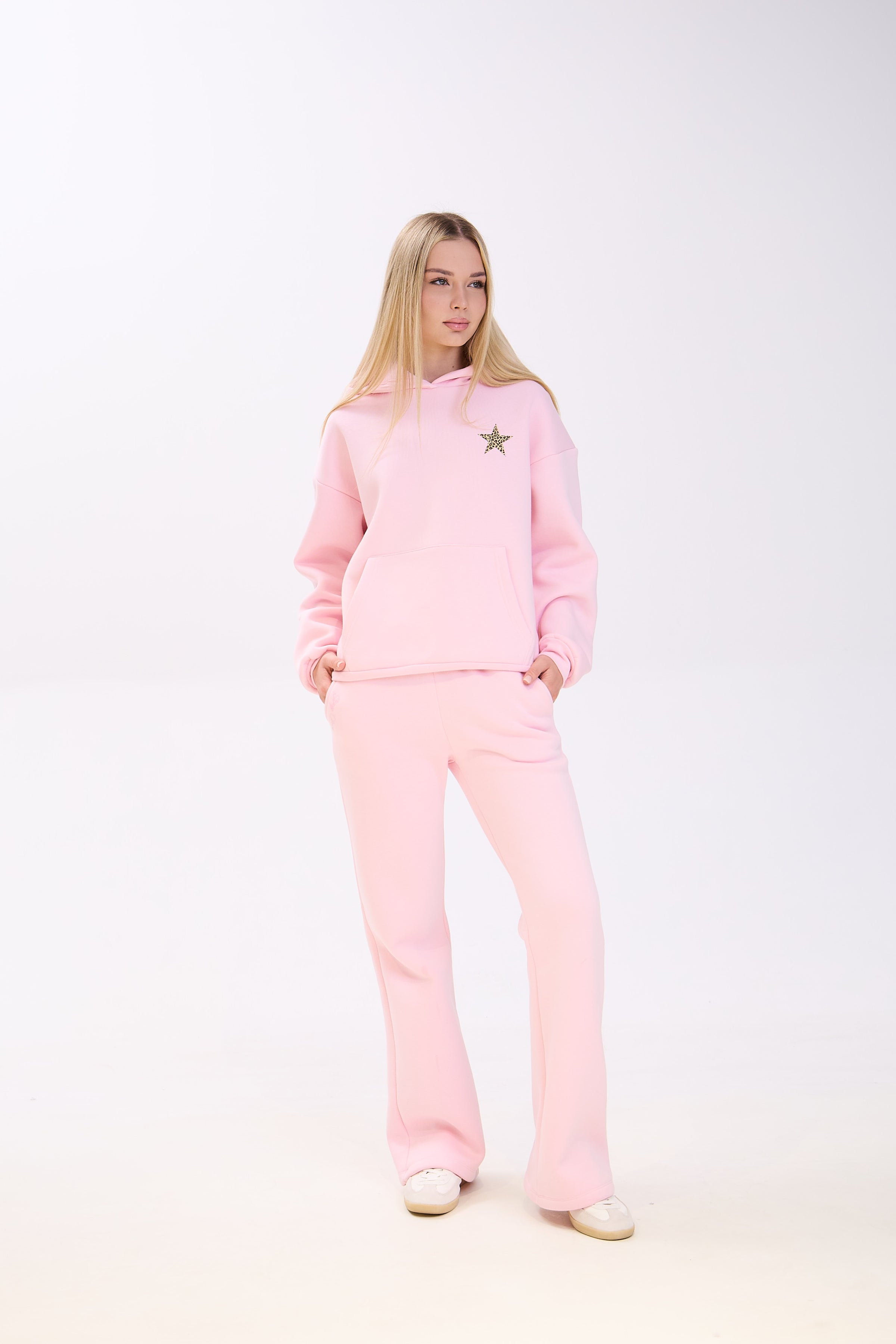 Woman dressed in a pastel pink hoodie with a leopard print star detail and matching pink wide-leg pants, posing with her hands in the pockets against a white studio background.