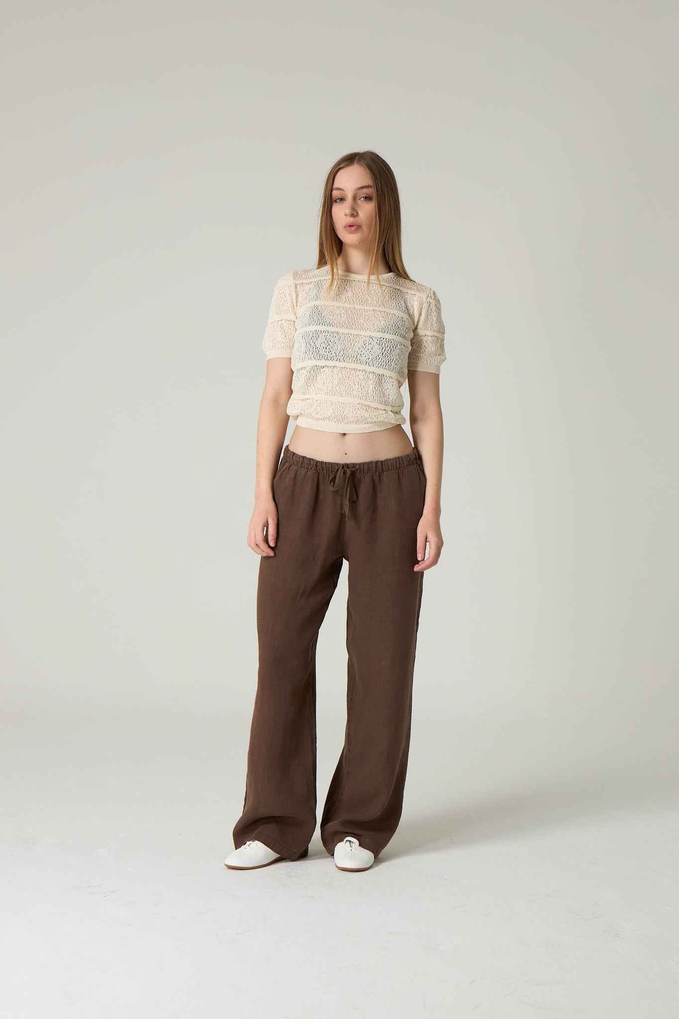 Woman wearing a sheer top and brown pants on a plain background.